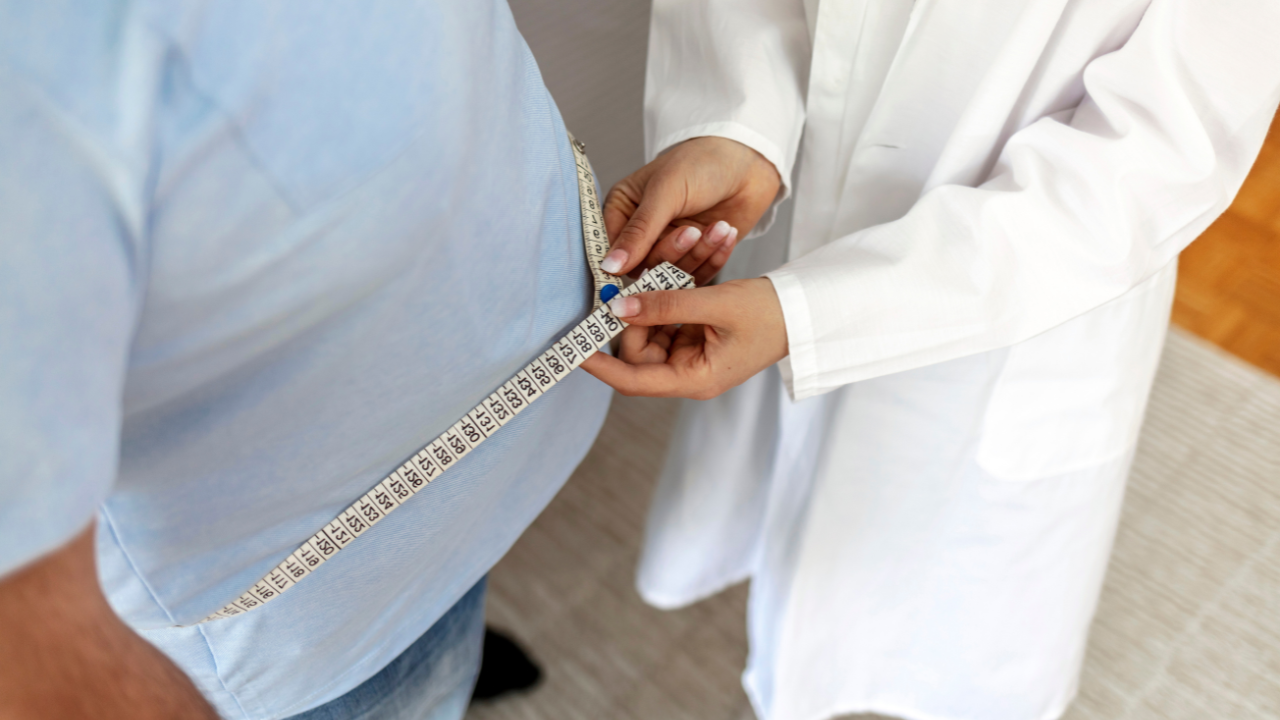 Women doctor measuring overweight man in clinic. Young female doctor measuring fat layer of overweight man with tailor tape in clinic. Eating, diet and unhealthy life concept. Copy space. Image Credit: Adobe Stock Images/Jelena Stanojkovic