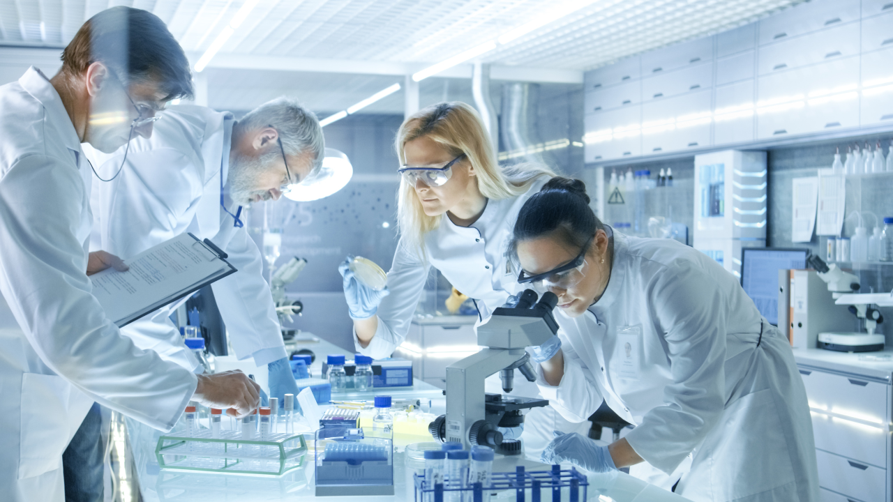 Team of Medical Research Scientists Work on a New Generation Disease Cure. They use Microscope, Test Tubes, Micropipette and Writing Down Analysis Results. Laboratory Looks Busy, Bright and Modern. Image Credit: Adobe Stock Images/Gorodenkoff